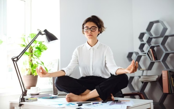 Seorang wanita bermeditasi di atas meja kantor (Foto: Freepik/cookie_studio)